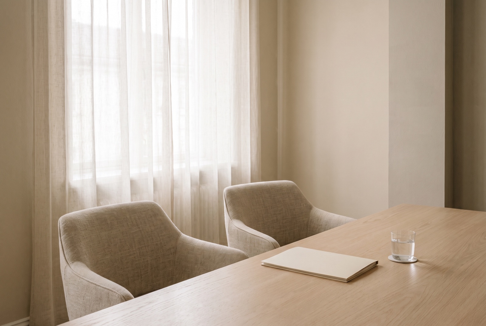 Two empty linen chairs at a pale oak meeting table by a sheer-curtained window, a closed folder and a glass of water