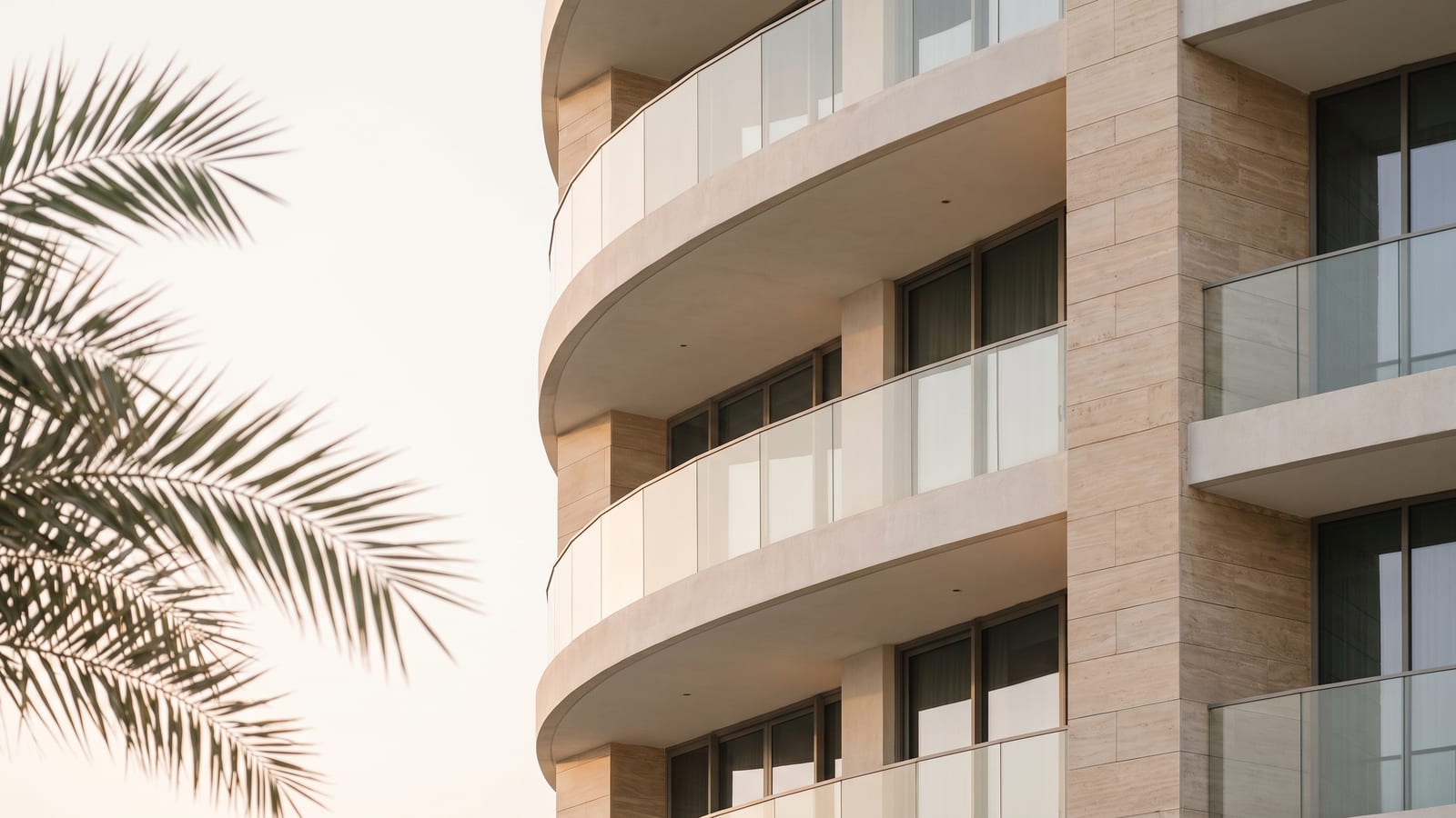 A curved contemporary residential facade with deep balconies, a palm frond across the frame